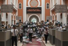 Un grand hall de bureau moderne au Maroc, avec des murs orange et des arches décoratives. Un tapis rouge s'étend au centre, menant à une montagne de dossiers papier et de boîtes qui bloque l'accès. Des employés vêtus de chemises blanches et de tenues traditionnelles naviguent, certains au téléphone, semblant accablés par la paperasse. Des panneaux directionnels bilingues sont visibles. L'ambiance est celle d'une inertie et d'une surcharge bureaucratique.