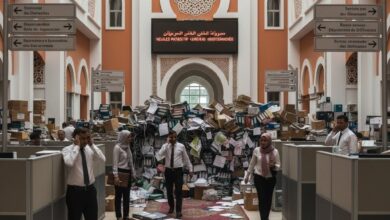 Un grand hall de bureau moderne au Maroc, avec des murs orange et des arches décoratives. Un tapis rouge s'étend au centre, menant à une montagne de dossiers papier et de boîtes qui bloque l'accès. Des employés vêtus de chemises blanches et de tenues traditionnelles naviguent, certains au téléphone, semblant accablés par la paperasse. Des panneaux directionnels bilingues sont visibles. L'ambiance est celle d'une inertie et d'une surcharge bureaucratique.